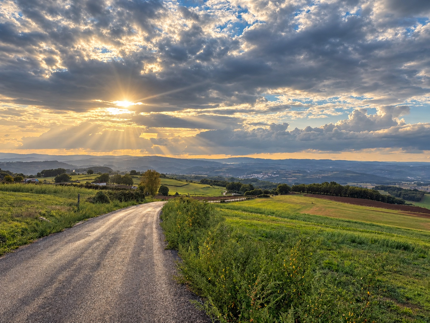 Carretera entre campos verdes bajo un cielo de nubes atravesado por rayos de sol
